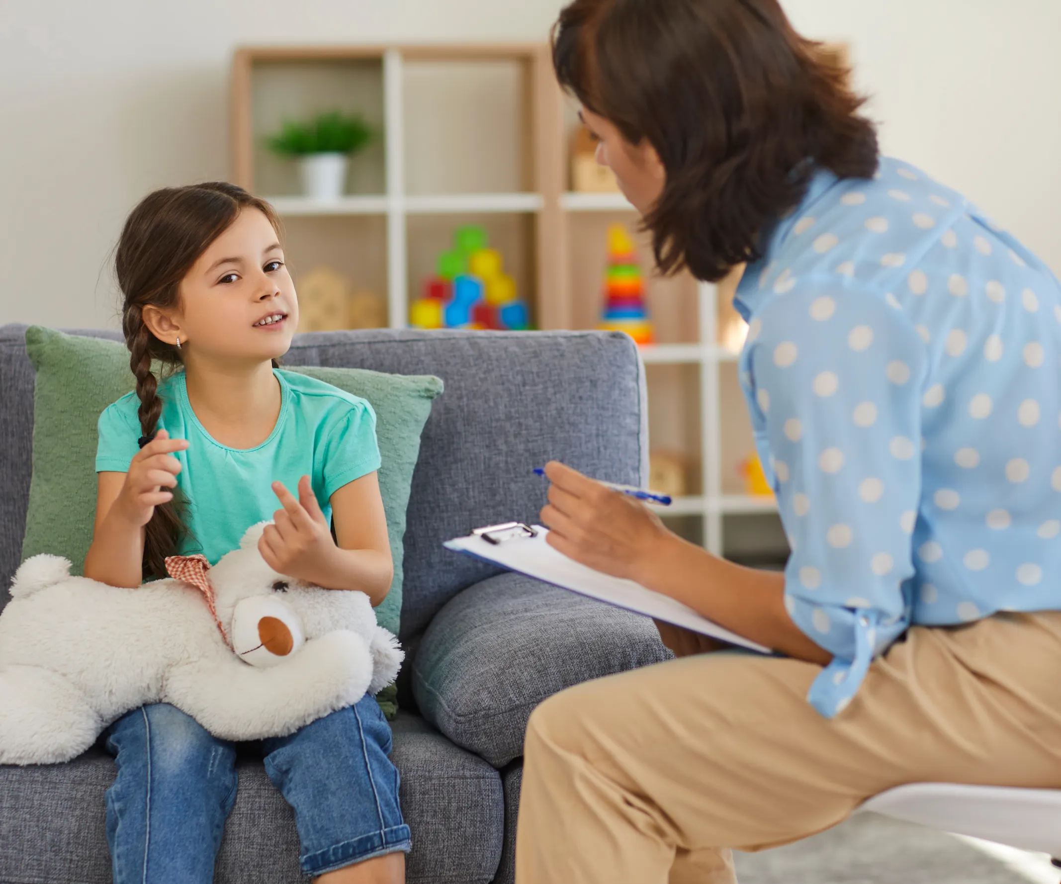 A girl holding a teddy bear sits on a couch, talking to a woman with a notepad.